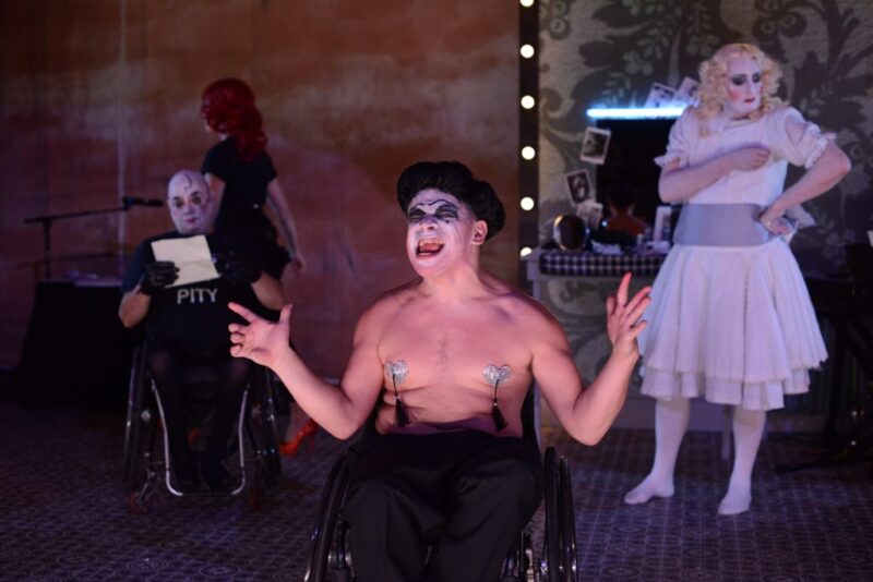 A shirtless man with a painted face wearing nipple tassles and a hat sits at the front of the stage and scowls. He is white and sat in a wheelchair .There are two more actors in drag makeup placed behind him..