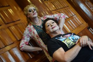 A close up of 2 drag queens. One has short blonde hair and the other has short black hair. 