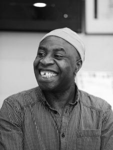 A black and white photo of Tayo Akinbode. He has a big smile and is wearing a beanie. 
