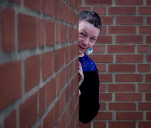 A photo of Caroline Parker. She has short dark hair and is peering out playfully from behind a red brick wall.