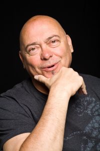 A photo of Garry Robson. He is bald, has blue eyes and is smiling with one hand to his face in a pose.