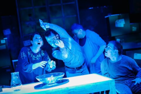Four people, two women and two men are gathered around a table, the stage is washed a mysterious blue. On the table there is a glass jug and a glass bowl. One of the men is standing over the glass bowl with something in his hands. The others are looking on in amazement.
