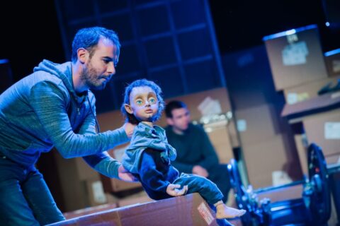 A man leans over a cardboard box that he has sat a puppet on, The puppet is of a small boy, he is sat upright and is looking out. He has large blue eyes, messy hair and appears to be wearing a blue tracksuit.