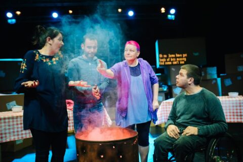 Four people, two men and two women are gathered around a rusty barrel that appears to have a fire in it. One of the men is holding his hands above it, as if to warm them. The others appear to be conversing. One woman is holding her arms out in a shrug, while the other woman is reaching out her arm and shaking a finger, as if in disapproval.