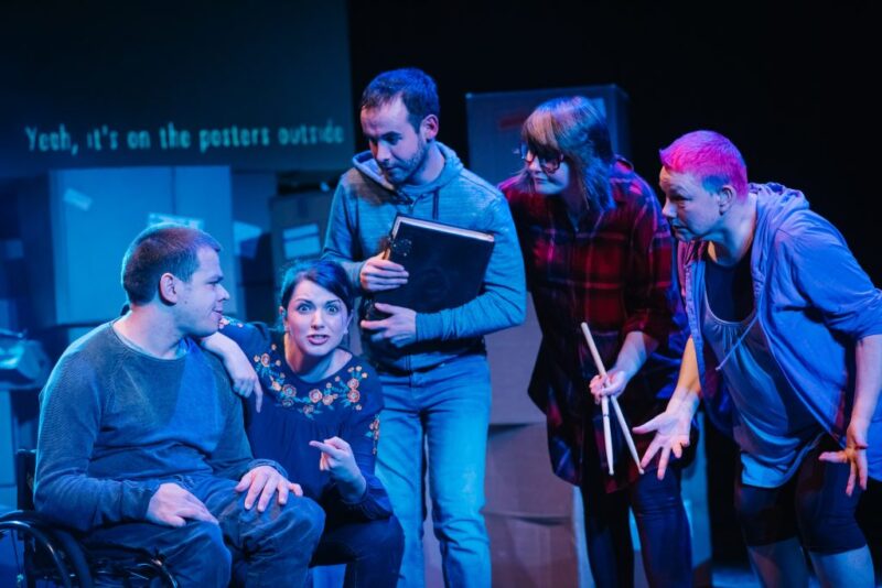 A group of people gather and lean in to listen to a woman talk. She appears to be in her early 30s and has dark hair. She has one of her arms leant on a man's shoulder, the other four people appear to be listening intently . They are all in modern dress, one of the men appears to be holding a large book and one of the women is holding drum sticks.