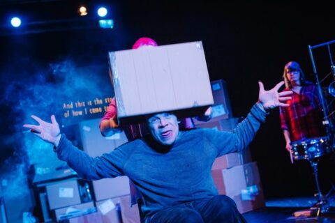 A woman places a cardboard box on a man in a wheelchairs head. He has his arms out wide and his tongue is stuck out. He is appears to be in his 30s and is wearing a blue jumper and blue trousers.