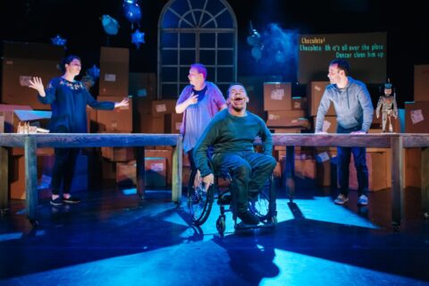 In this scene, the background is set with a large glass window with star shaped balloons around it. In front of this there is a wall of cardboard boxes and in front of them there are two large wooden tables. On one of the tables, the toy soldier is stood. Two women and a man are stood spread out behind the tables. The man is leaning with both hands on the table in front of him. The woman in the middle is pointing to herself while looking to the other woman who is throwing her arms up at her sides. A man in a wheelchair moves in front of the tables with a big grin on his face which is covered in chocolate.