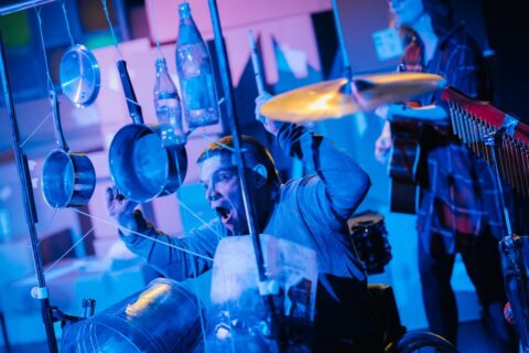 A man sits behind a large metal frame that has various things suspended from it with wires. There are pots, pans, glass bottles and buckets. The man's eyes and mouth are wide open. He is holding a drumstick in each hand and has his arms thrown back as if about to starts hitting the items. Behind him there appears to be a woman playing guitar.