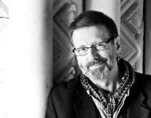 A black and white photo of Mike Kenny. He has a greying beard, thin framed glasses and is wearing a scarf. 