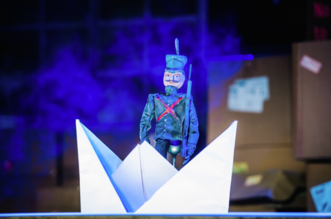 A small soldier figurine sits in a white paper boat. He is in a green regency style soldier uniform with a red cross at the front. He has a curly mustache and appears to have one leg missing.
