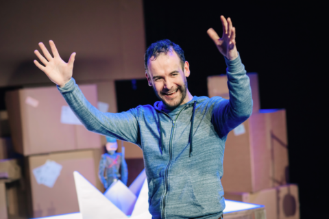 A photo of a middle aged man with sparse curly dark hair. He is wearing a light blue hoodie and has his arms raised in the air. In the background there is a collection of cardboard boxes and the toy soldier sat in his paper boat.
