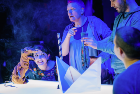 A table is lit up white, four people, two men and two women are gathered around it. On the table there is a white paper boat. One of the men is holding up a toy soldier in the boat. At the end of the table, a young woman is crouched down with a brown puppet of a scraggly looking rat. In the middle of them there is a middle aged woman with pink hair who appears to be signing.