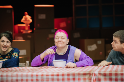 Three people, two women and a man, sit at a long dinner table that is covered with a red and white checked table cloth. They each have a white bowl in front of them. The woman in the middle is lifting a spoon to her face which has a wide grin on it. She has short bright pink hair and appears to be in her late 40s. To the right there is a younger woman in a blue shirt who appears to be talking, a man on the other side appears to be listening to her as he leans forwards.