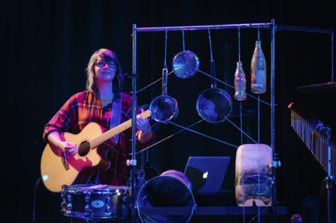 A young woman in a red checkered shirt stands playing guitar. In front of her there is a small drum, and next to that is a metal frame that has cooking pots and empty bottles hanging from it.