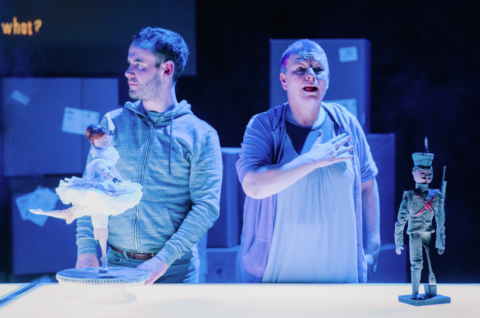 A man and a woman stand behind a bright white table with a toy soldier and a toy ballerina on it. The woman has very short hair, she appears to be in her late 40s and she is holding one of her hands over her heart. The man appears to be in his 30s, he is wearing a blue hoodie and looking off to the side at something out of frame.