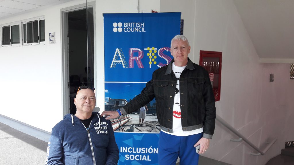 A photograph of Garry Robson and Kinny Gardner. They are in front of a banner that says 'British Council: Arts'. Garry is a white, middle aged bald man in a wheelchair. He has a pair of sunglasses on his head and is wearing a blue zipped up hoodie. Kerry also appears to be white and middle aged. He has short white hair. He is wearing blue trousers, a white T shirt with a face printed on it and a dark blue denim jacket. 