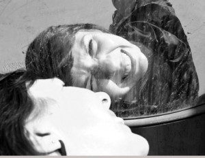 A woman with short hair and gritted teeth turns her head to one side and looks at her reflection. The photo is taken from such an angle as we see more of the reflection that her. 