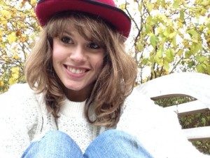 A photo of Lynn Pilkington. She is wearing blue jeans, a white top and a red hat. She has medium length brown hair and has a big smile. 