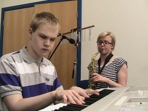 A photo of Matthew Ward playing a keyboard. There is a girl sat beside him playing the saxophone. 
