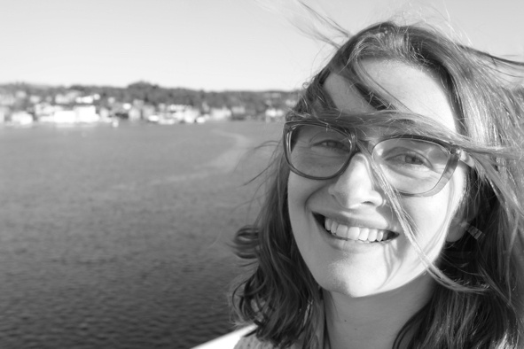 A black and white photo of Mairi Taylor that look to have been taken on a ferry. She appears to be in her mid 30s, white, she has medium length dark hair and glasses. She is smiling at the camera.