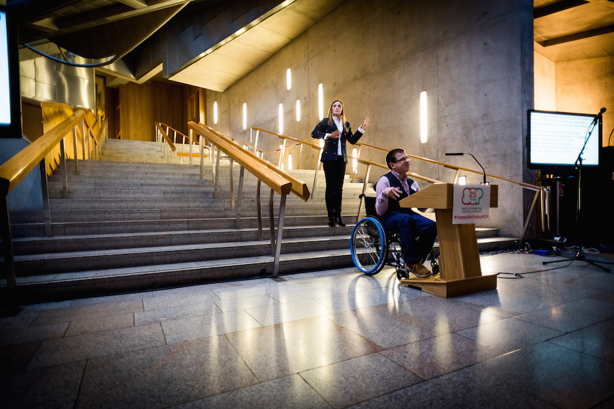 Robert talks in front of a wide, shiney set of stairs. He is sat in a wheelchair behind a small wooden podium that displays the Birds of Paradise logo. He has short brown hair and is wearing a smart shirt with a waistcoat. Behind him a woman appears to be interpreting. She is wearing a suit and has long blonde hair.