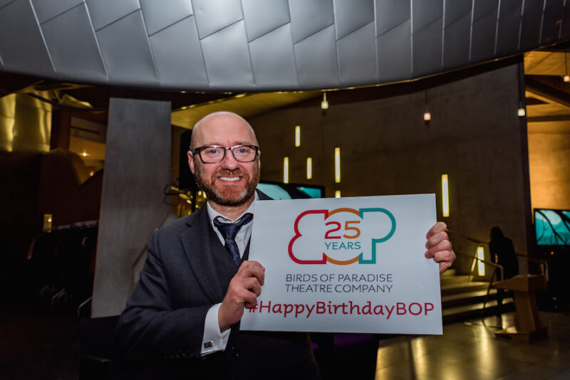 A man smiles holding a sign with the Birds of Paradise logo with '25 years' written inside it. Underneath this it says #HappyBirthdayBOP. The man appears to be in his late 40s, white, he is bald with a brown beard, is wearing glasses and a grey three piece suit.