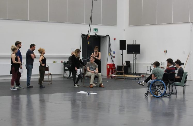 Image from rehearsing a scene: In a large white and grey room with no set, one person sits in a chair while two people fuss aorund him and the rest of the cast stand in a line and watch. Watching them are Robert, Scott and EJ - who are the creative team.