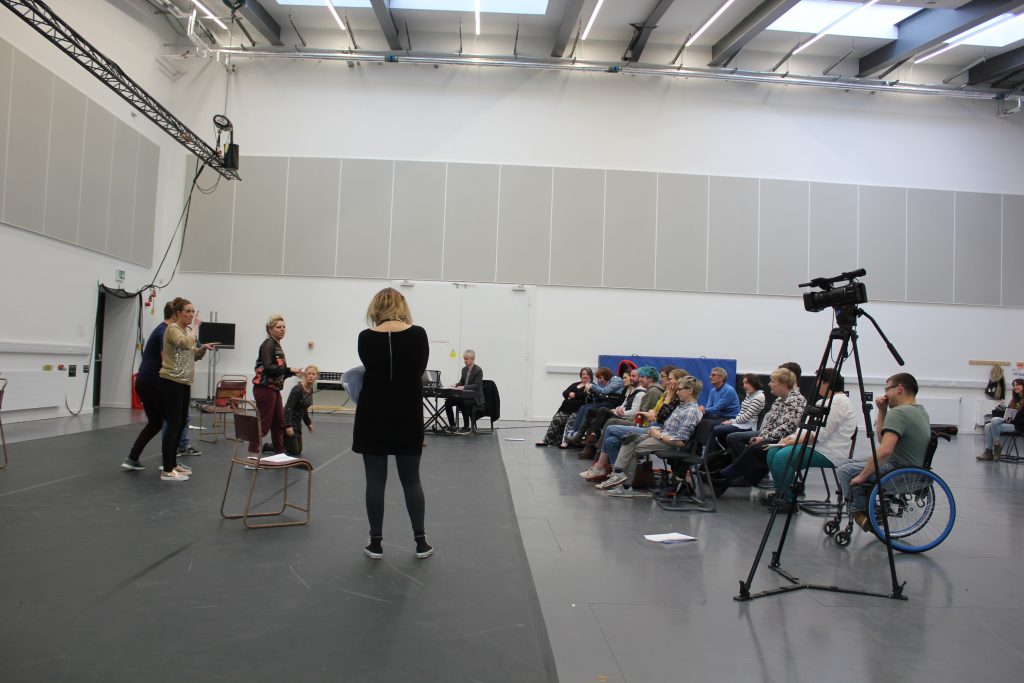 Side view of the sharing: On the right, the cast go through the scenes they have rehearsed - there are no costumes or set and their scripts are scattered about close to hand, on the right are two rows of an invited audience from National Theatre of Scotland.