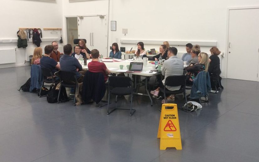 Distance shot of the first read through: everyone is sitting round a large table covered in papers, water and laptops.