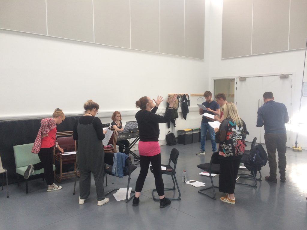 Process of working on the script: The cats stand in a semi circle around Claire sitting at a keyboard. They each have their scripts in their hands apart from Nat who is practicing the signing.