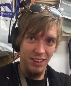 A photo of Nic Stuart. He has messy brown hair, a lip piercing and is wearing sunglasses on top of his head.