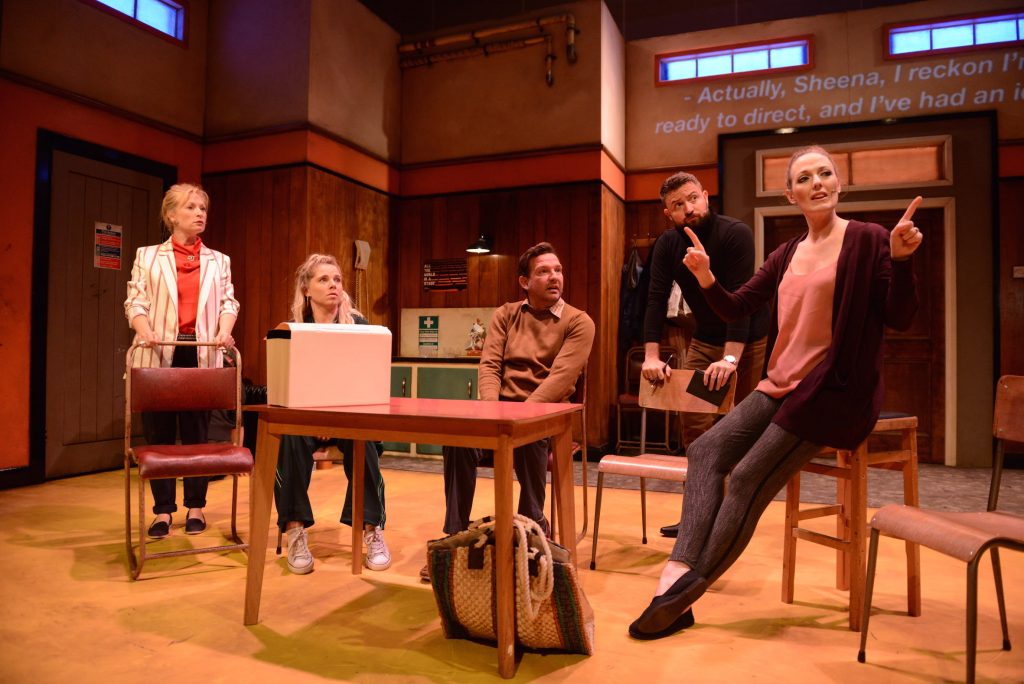 5 actors are sat around a desk on stage. They all look to an actor on the right who appears to be explaining something.