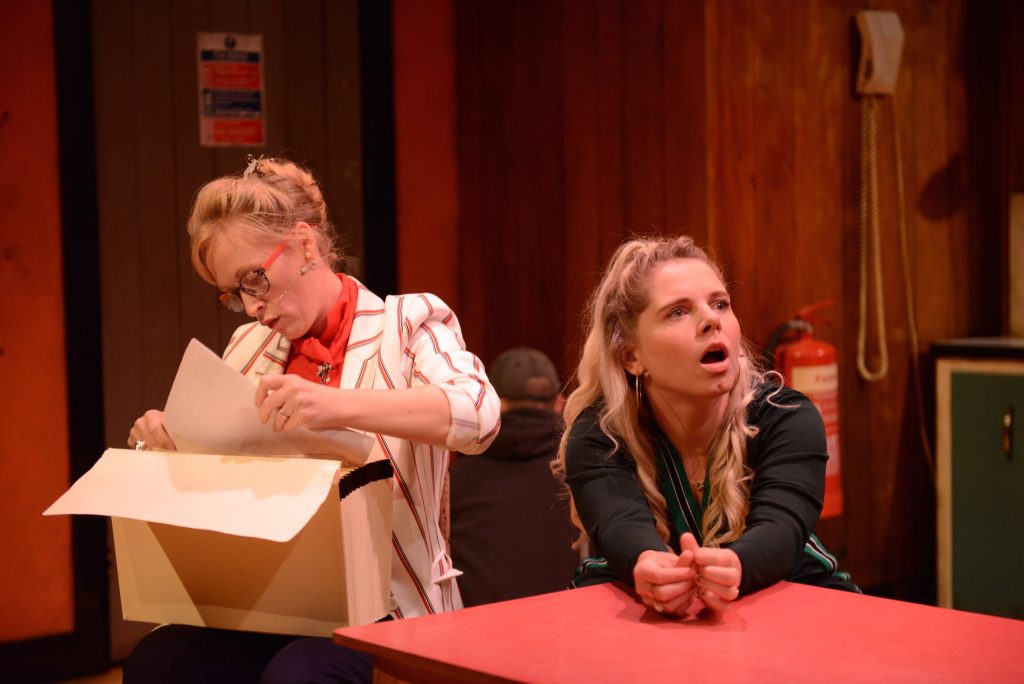 2 actors are on stage. One of them is looking through a box of things, and the other is looking at something outside of the photo with a shocked expression on her face