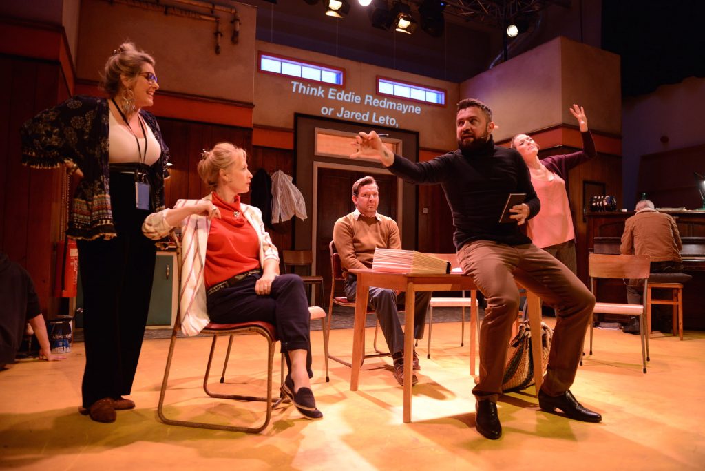 A group of 5 actors gathered around a desk. One of them is sitting on the desk and appears to be explaining something while the other look on.