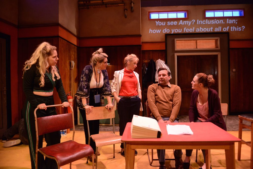 5 actors are sat round a desk. They have all turned to a man in the middle who appears to be telling them about something.