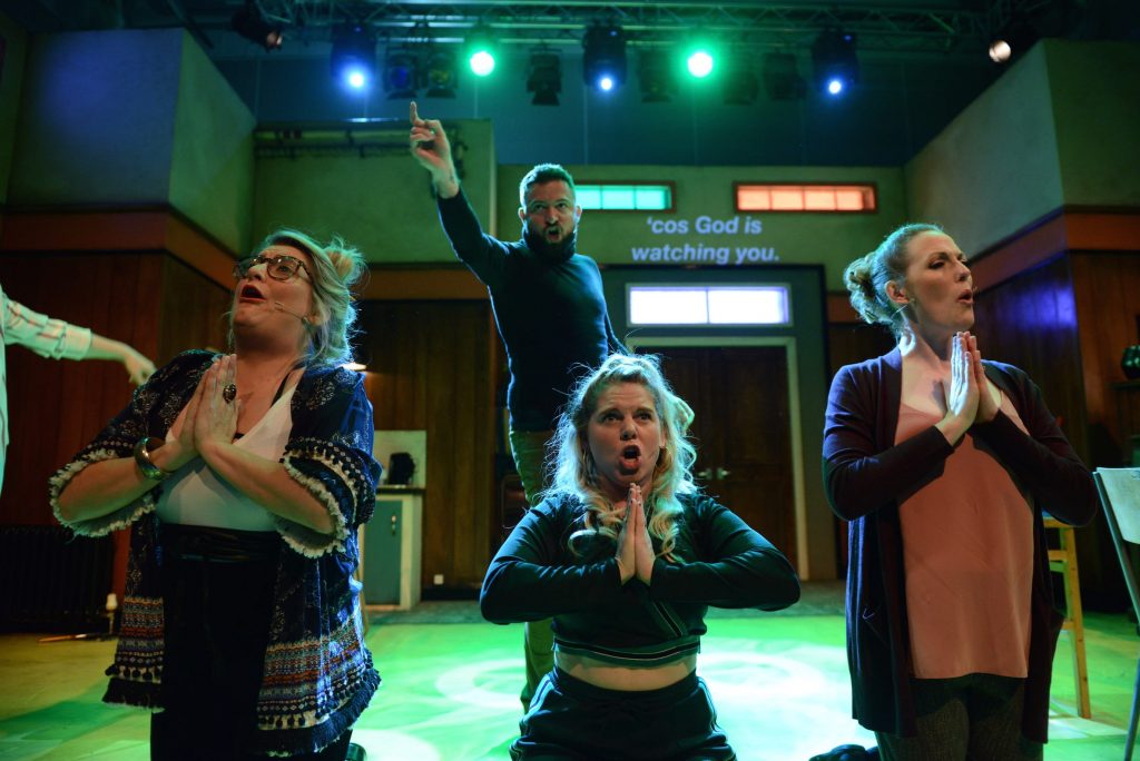 3 actors kneel at the front of the stage, with their hands together as if they are praying. A man stands behind them and points up to the air above him.