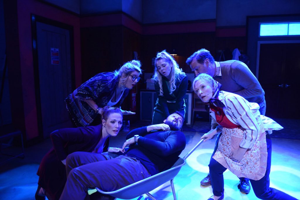 6 actors huddle at the front of the stage which is lit blue. One of the actors lies in a wheelbarrow which the others are pushing.