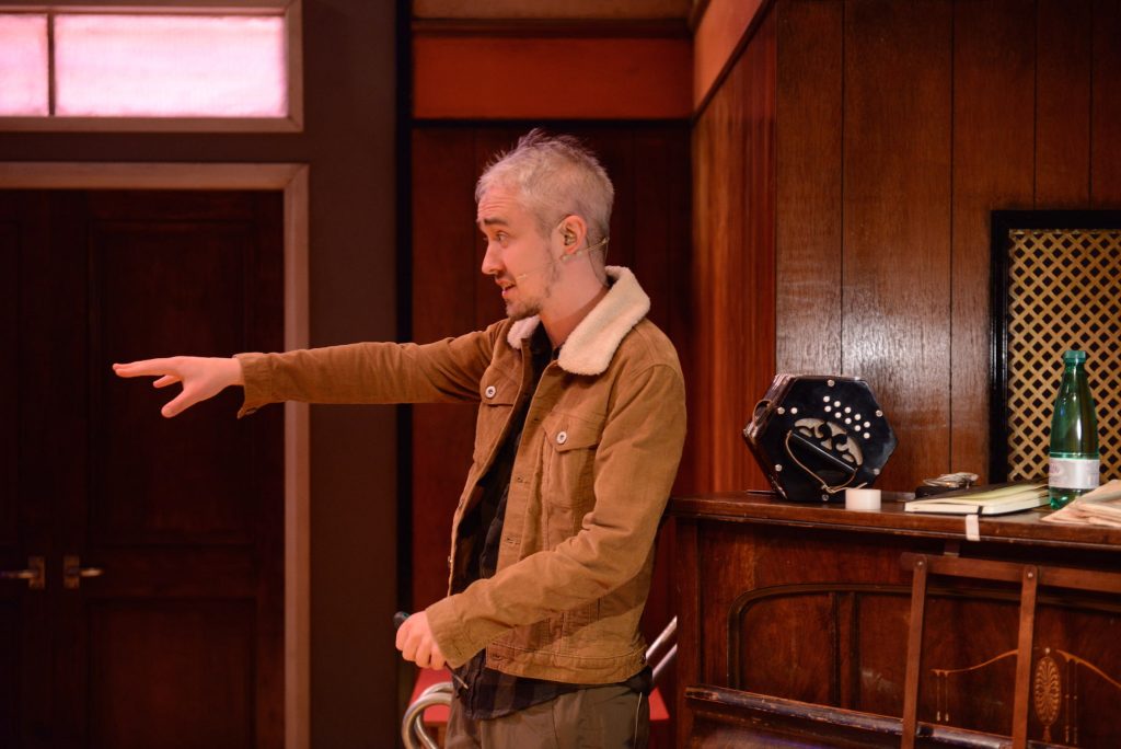 A man with short grey hair, wearing a brown jacket, gesticulates to something outside of the photo.