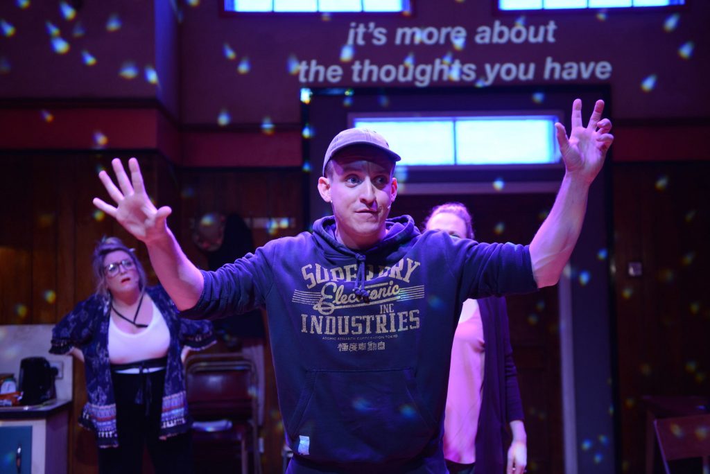 A man wearing a hoodie and a cap stands at the front of the stage with his arms in the air. Two actors behind him look on.
