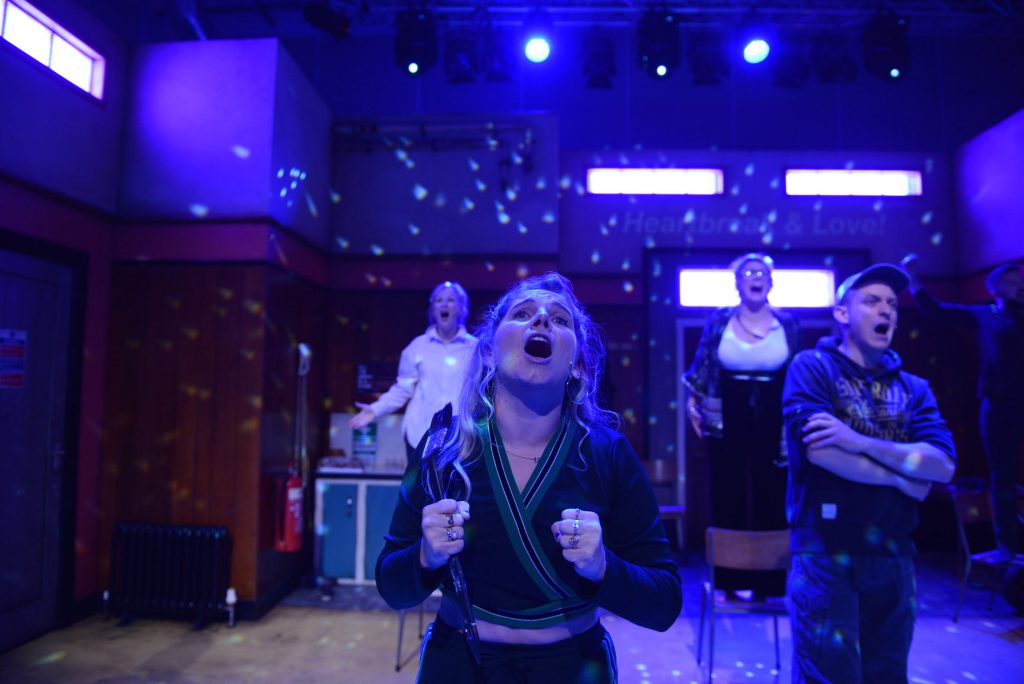 A close up of four actors singing on stage. The stage is lit with a blue light with white dots.