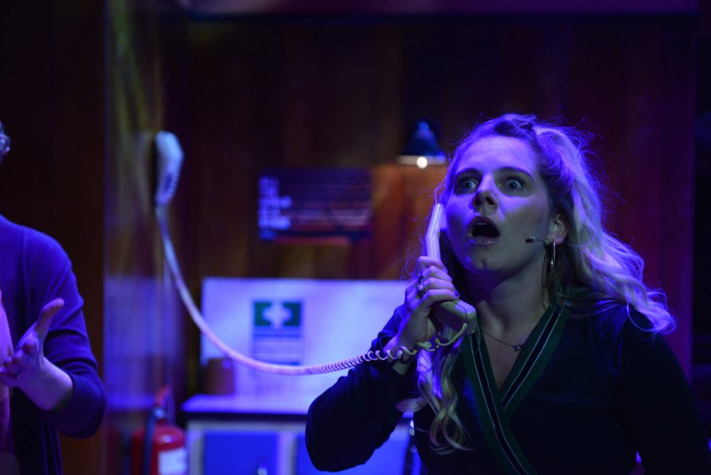 An actor on stage on the phone. She looks shocked at what she is hearing.