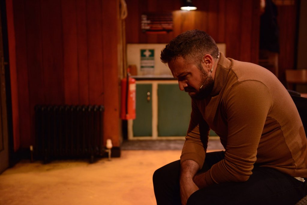 An actor in a brown jumper sits hunched over on stage. He appears upset.