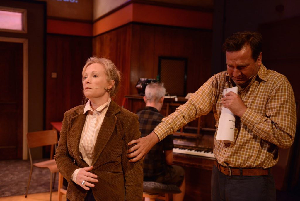 Two actors appear at the front of the stage. One of them appears to be crying, he has his hand reached out onto the other actors arm.