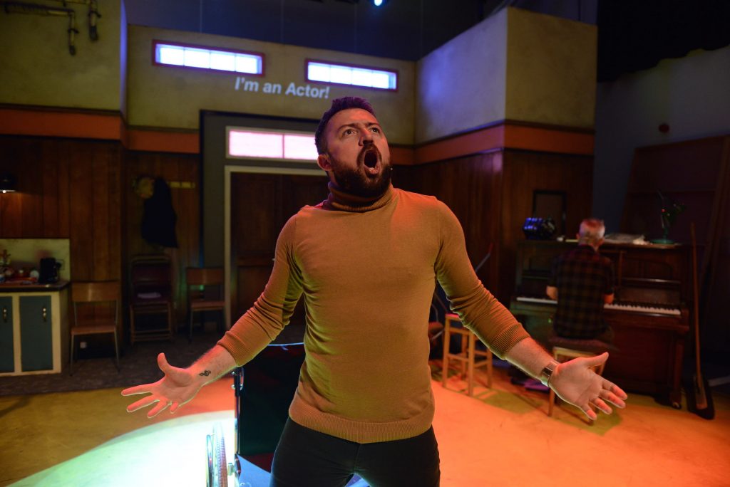 An actors is singing at the front of the stage his mouth and his arms are open wide.