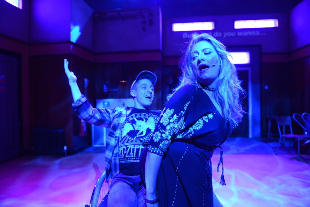 An actor with a cap on sits in a wheelchair. A woman is crouching in his lap. They both appear to be singing.