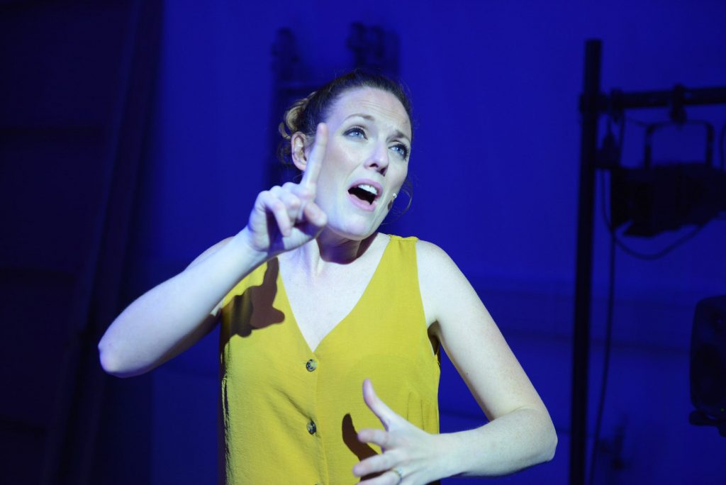 A close up of an actress in a yellow top on stage. She appears to be singing and signing.