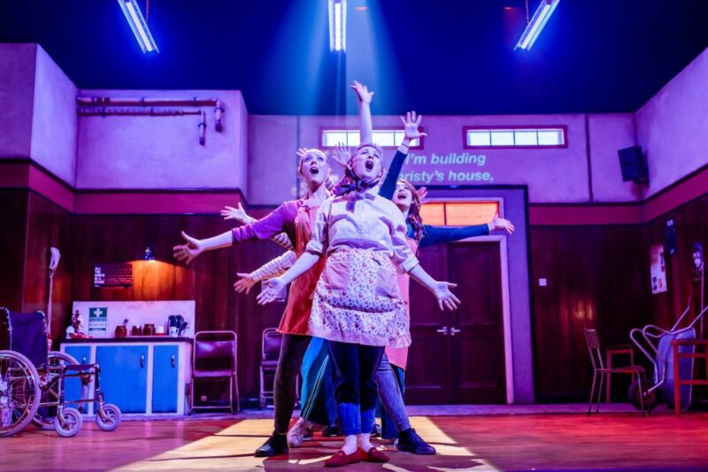 Five actors line up behind each other centre stage. They have their arms outstretched and the faces that we can see have their mouths open wide as if they are singing.