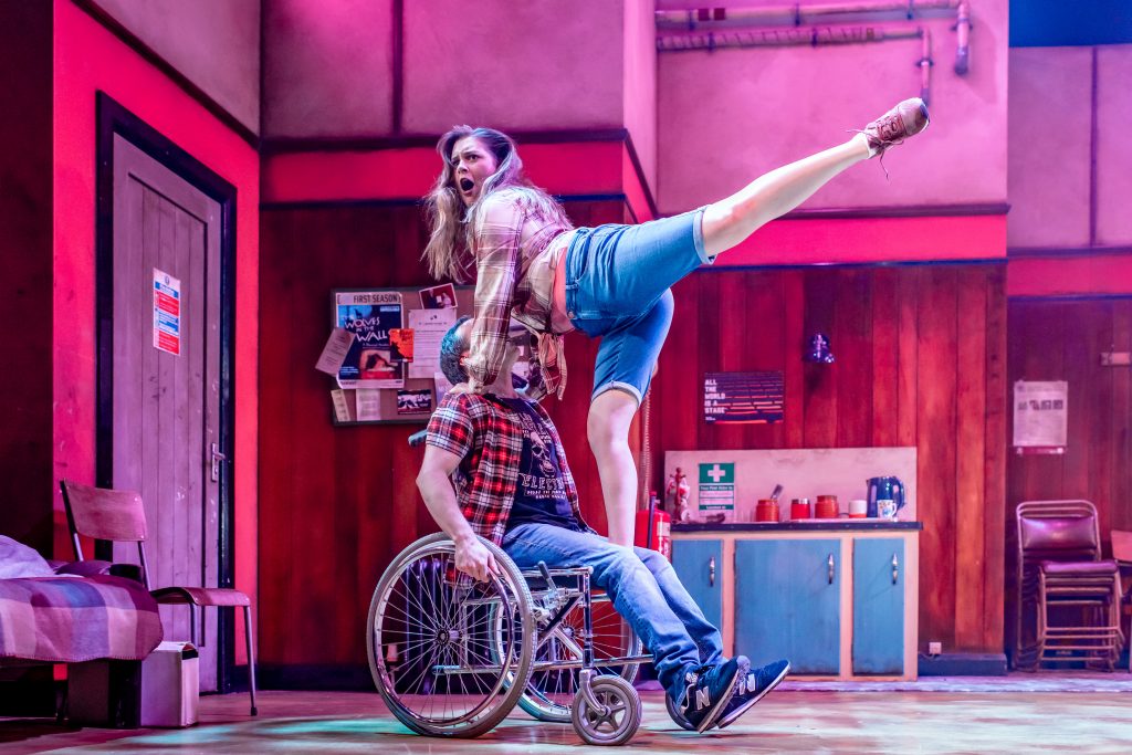 A woman stands on a mans wheelchair. She has one foot on the chair and the other is up in the air. Her hands are on the man's shoulders for support. She has long blonde hair, and is wearing blue shorts and a plaid shirt. The man, whose face is slightly obstructed, is wearing blue jeans, a black T shirt and a plaid shirt.