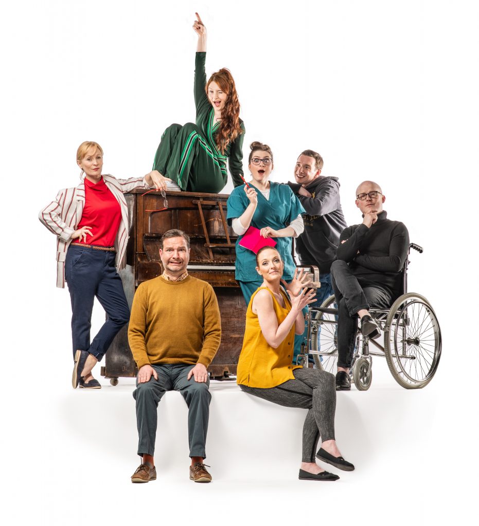 A poster for the show. It features seven cast members, they are all gathered around a large piano, with one woman sat on top of it. They are all in modern clothes and smiling at the camera