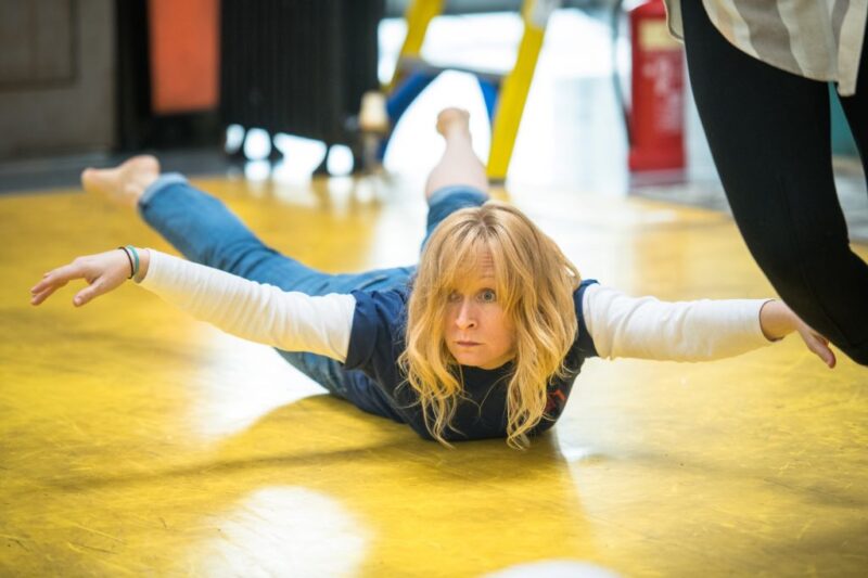 A middle aged woman lies stomach down on a yellow floor. She has her arms and legs lifted off the floor, as if pretending to fly. She is wearing blue jeans and a blue top and has medium length blonde hair.